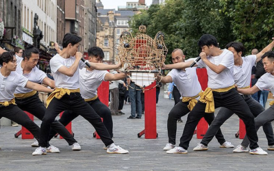 A group of nine people in matching white shirts and yellow sashes pose in a coordinated martial arts stance on a city street, holding a decorative structure in the centre, with buildings and trees in the background.