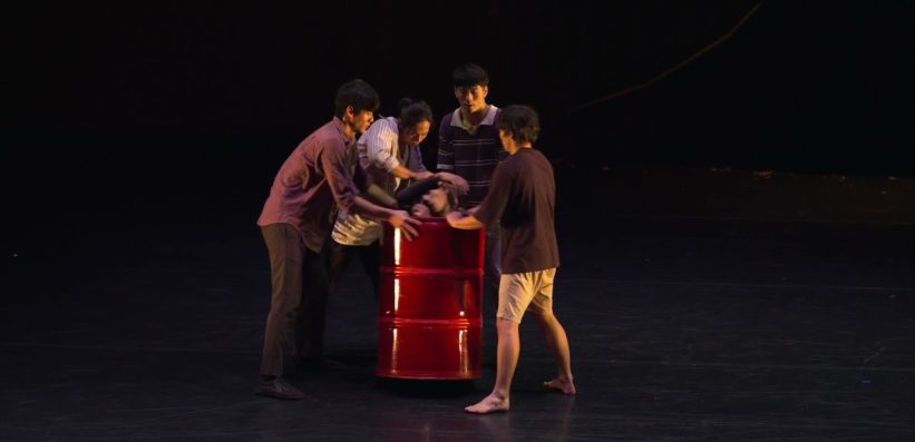 Five people stand on a dark stage, surrounding and holding a large red barrel. One person is partly inside the barrel with only their head and arms visible, whilst the others interact and support the barrel.