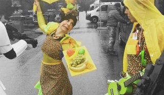 Two people in colourful costumes stand outside; one in a fruit-themed outfit with a tray of fake fruit and a yellow umbrella, the other with a green guitar, rain mac, and yellow hat. The background is mostly greyscale.