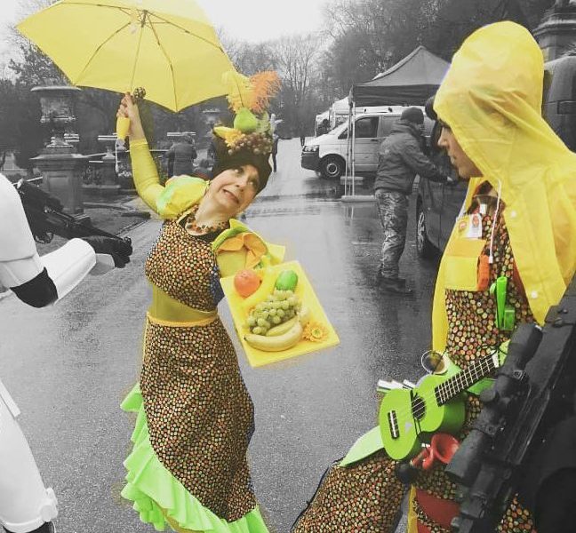 Two people in colourful costumes stand outside; one in a fruit-themed outfit with a tray of fake fruit and a yellow umbrella, the other with a green guitar, rain mac, and yellow hat. The background is mostly greyscale.