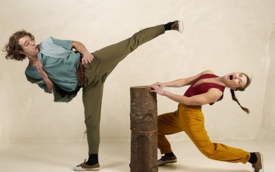 Two people pose comically: one with curly hair, in green trousers, performs a high kick over a tree stump, while the other in yellow trousers leans back dramatically, making a surprised face. Both are indoors against a plain wall.