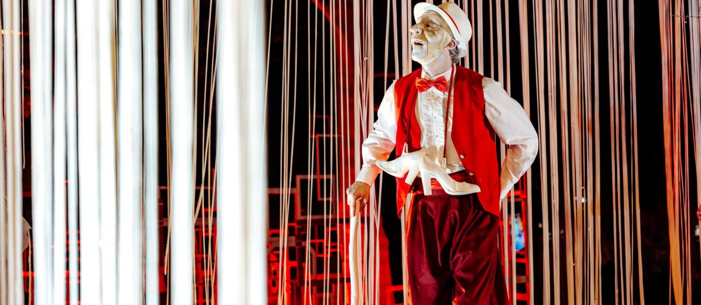 A performer dressed in a red waistcoat, red trousers, white shirt, and white hat stands among hanging white ribbons. His face is painted white, and he holds a white mask. The background is lit with red lights.