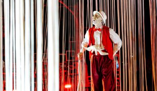A performer dressed in a red waistcoat, red trousers, white shirt, and white hat stands among hanging white ribbons. His face is painted white, and he holds a white mask. The background is lit with red lights.