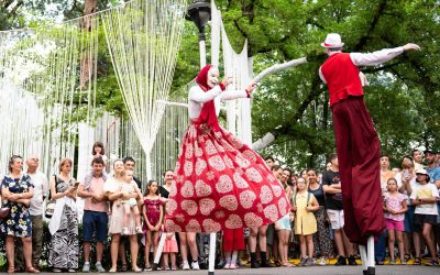 Two performers on stilts, dressed in colourful costumes, entertain a crowd of people outdoors. The crowd, including children and adults, watches eagerly. Trees and decorative white streamers are visible in the background.