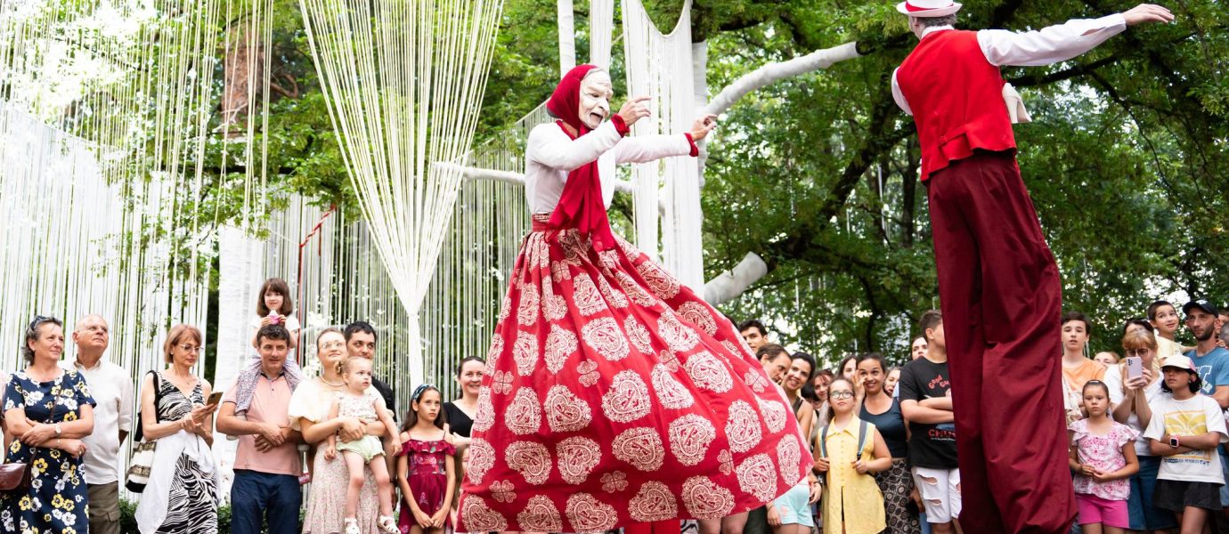 Two performers on stilts, dressed in colourful costumes, entertain a crowd of people outdoors. The audience, including children and adults, watches with interest, surrounded by trees and decorative white streamers.