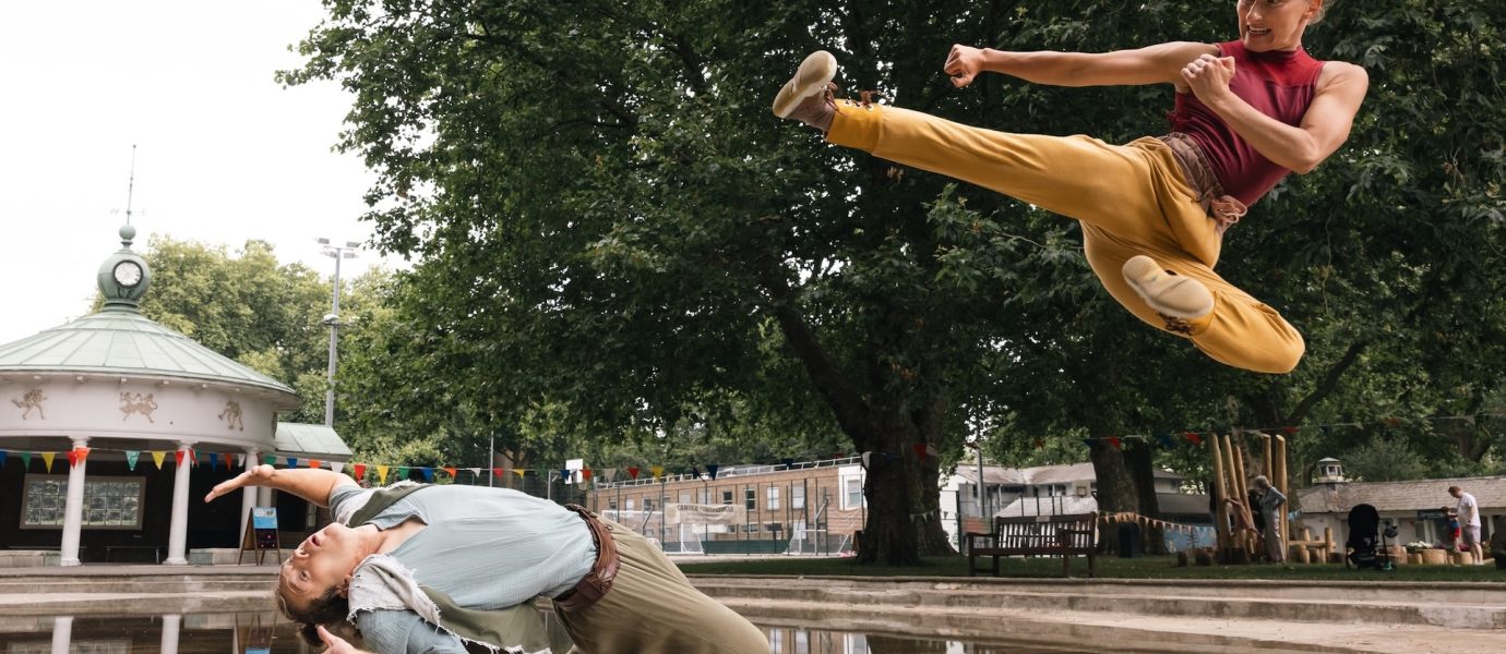 Two people perform martial arts outdoors; one person is mid-air doing a high kick while the other leans backwards dramatically, dodging the kick, with trees and a pavilion in the background.