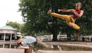 Two people perform martial arts outdoors; one person is mid-air doing a high kick while the other leans backwards dramatically, dodging the kick, with trees and a pavilion in the background.