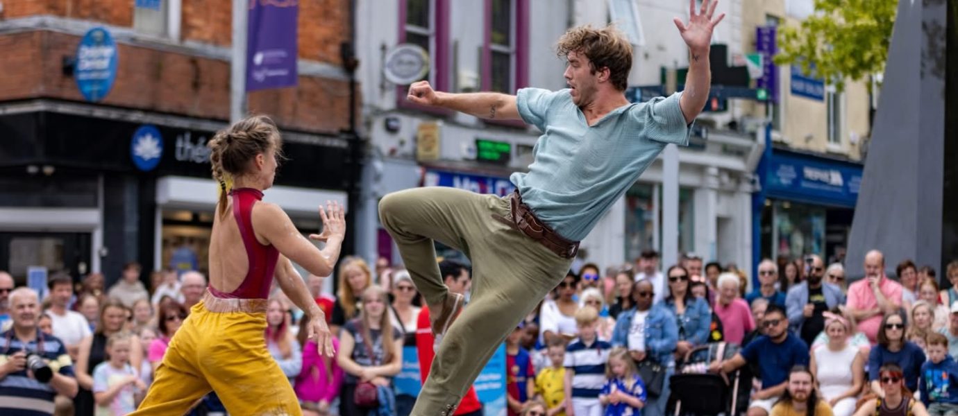 Two street performers entertain a crowd; one woman in yellow trousers faces a man in mid-air with a raised leg and arm, as onlookers watch in a lively city scene. The Waterford logo is visible in the background.