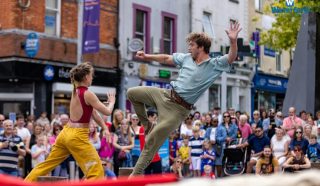 Two street performers entertain a crowd; one woman in yellow trousers faces a man in mid-air with a raised leg and arm, as onlookers watch in a lively city scene. The Waterford logo is visible in the background.