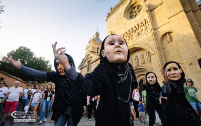 Performers wearing large, expressionless masks and black costumes interact with a crowd on a sunny day in front of an historic stone church, as spectators watch and take photos.