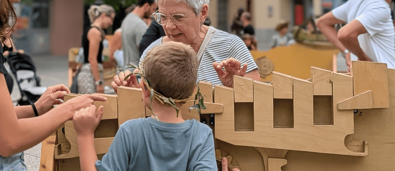 An older woman and a child play with a large wooden mechanical puzzle outdoors, surrounded by other people. The child wears a grey shirt and a leafy headband. The mood is lively and interactive.