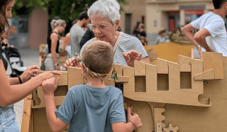 An older woman and a child play with a large wooden mechanical puzzle outdoors, surrounded by other people. The child wears a grey shirt and a leafy headband. The mood is lively and interactive.