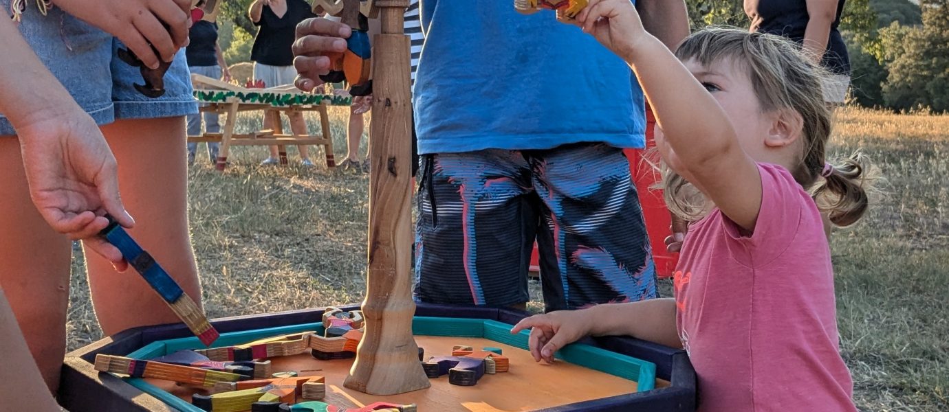 Several children are playing a tabletop game outdoors, stacking wooden animal pieces on a vertical pole. Adults and more children are visible in the background. The scene is set in a grassy area on a sunny day.