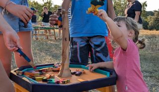 Several children are playing a tabletop game outdoors, stacking wooden animal pieces on a vertical pole. Adults and more children are visible in the background. The scene is set in a grassy area on a sunny day.