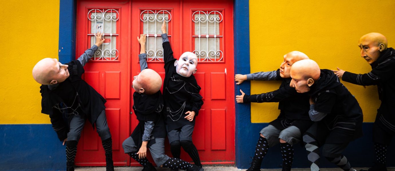Six people in theatrical costumes and large bald masks pose dramatically in front of a bright red door set in a yellow and blue wall on a cobbled street.