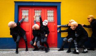 Six people in theatrical costumes and large bald masks pose dramatically in front of a bright red door set in a yellow and blue wall on a cobbled street.