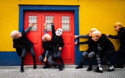 Six performers in black outfits and bald, exaggerated masks pose dramatically in front of a bright red double door set in a yellow wall with a blue border, creating a theatrical and surreal scene.