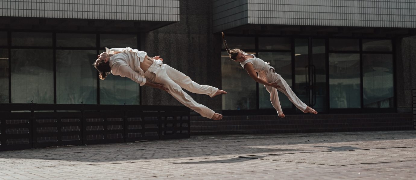 Two people dressed in white clothing are captured mid-air, parallel to the ground, as they perform synchronised jumps or acrobatic moves outside a modern building with tiled walls.
