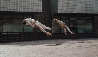Two people dressed in white clothing are captured mid-air, parallel to the ground, as they perform synchronised jumps or acrobatic moves outside a modern building with tiled walls.