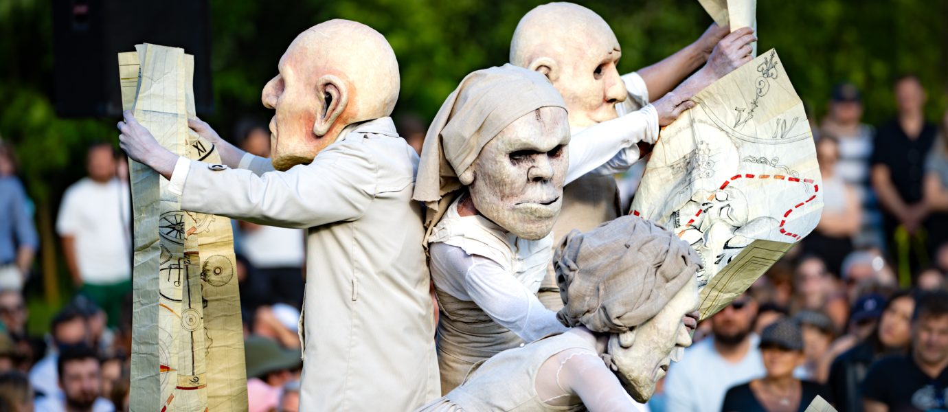 Four performers wearing large, exaggerated masks and costumes hold illustrated maps, entertaining a crowd at an outdoor event with trees and spectators visible in the background.