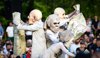 Four performers wearing large, exaggerated masks and costumes hold illustrated maps, entertaining a crowd at an outdoor event with trees and spectators visible in the background.