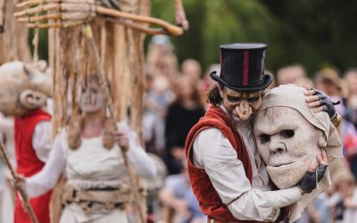 A performer in a red waistcoat and top hat hugs a large, white mask. Other performers in white costumes and masks hold puppets in the background. A crowd watches the outdoor theatrical event.