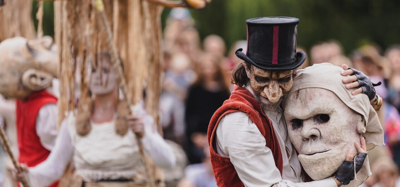 A performer in a red waistcoat and top hat, wearing a mask, hugs a large theatrical mask. Other masked performers in white costumes and a wooden puppet are visible in the background at an outdoor event.