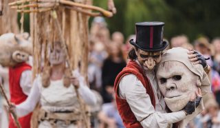 A performer in a red waistcoat and top hat, wearing a mask, hugs a large theatrical mask. Other masked performers in white costumes and a wooden puppet are visible in the background at an outdoor event.