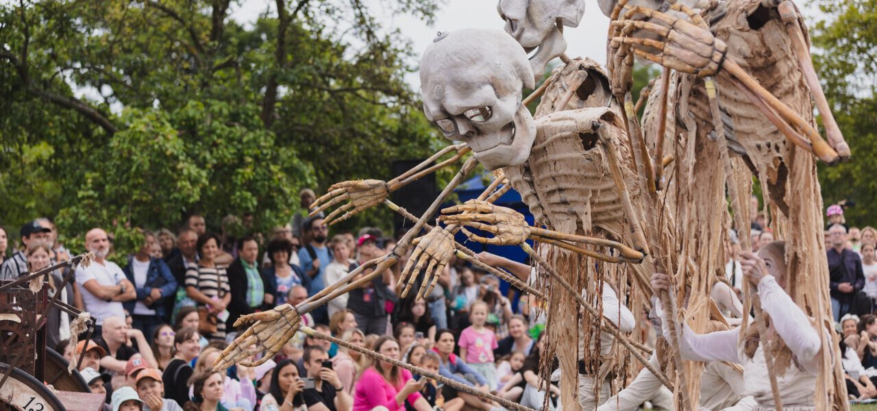 Three large skeleton puppets are operated by performers in white costumes in front of a seated crowd outdoors. Trees and greenery are in the background, and many people are watching the show.