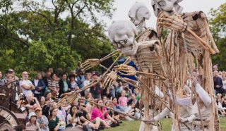 Three large skeleton puppets are operated by performers in white costumes in front of a seated crowd outdoors. Trees and greenery are in the background, and many people are watching the show.
