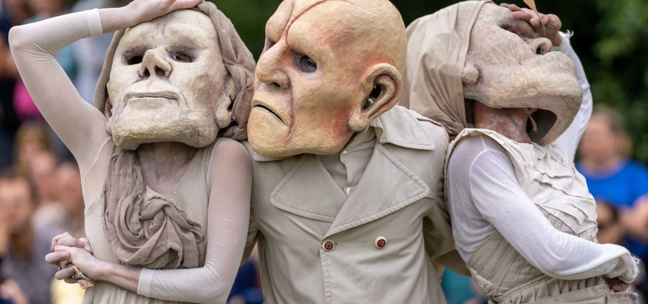 Three performers wearing large, exaggerated masks and beige costumes pose dramatically outdoors, with an audience and greenery blurred in the background.