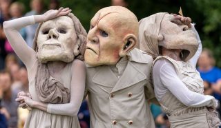 Three performers wearing large, exaggerated masks and beige costumes pose dramatically outdoors, with an audience and greenery blurred in the background.