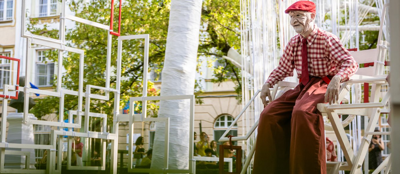 An elderly person in a red chequered shirt, red hat, and wide red trousers sits on a white chair amid an outdoor art installation of white and red geometric frames, with trees and buildings in the background.