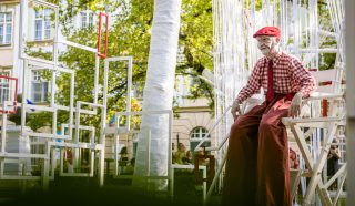 An elderly person in a red chequered shirt, red hat, and wide red trousers sits on a white chair amid an outdoor art installation of white and red geometric frames, with trees and buildings in the background.