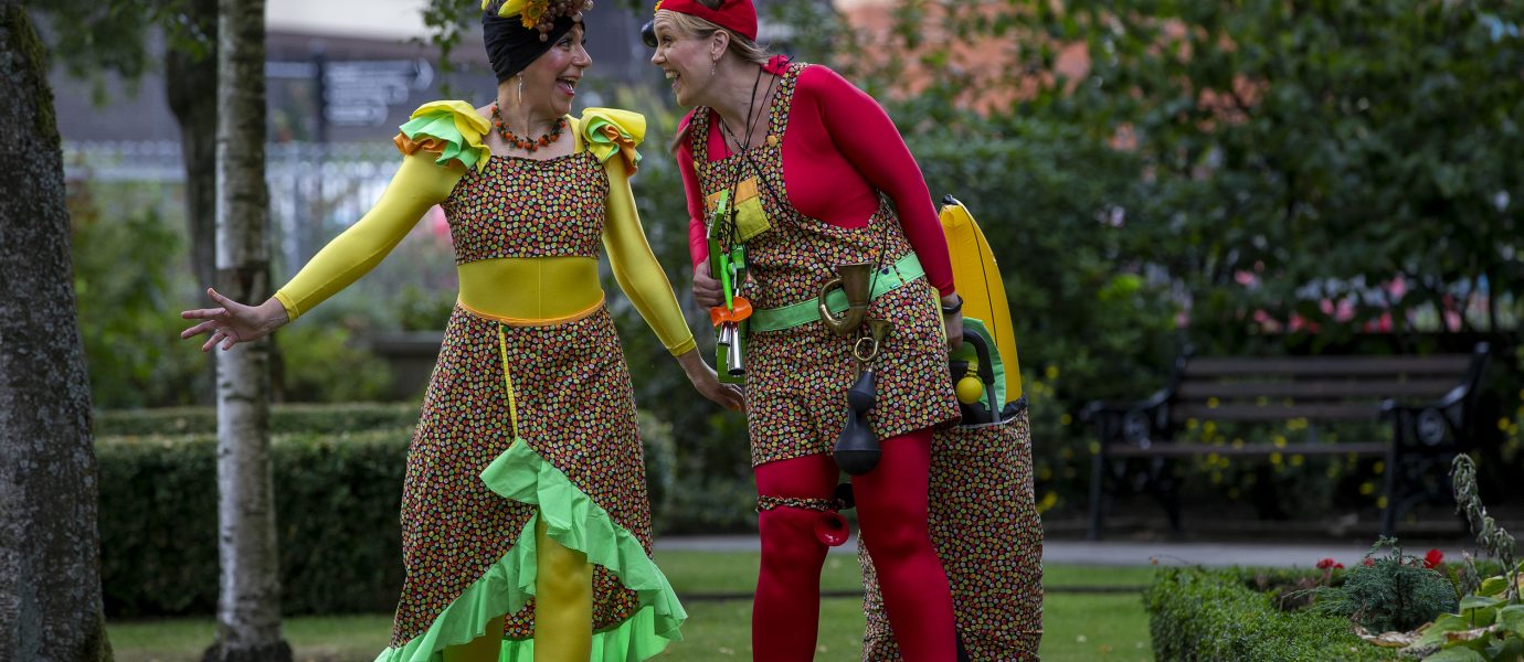Two people in colourful costumes and hats, smiling and interacting in a garden park. One wears yellow and green, the other red, both with floral patterns, and one has a small trolley decorated with flowers.