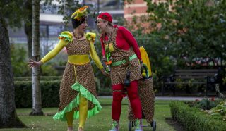 Two people in colourful costumes and hats, smiling and interacting in a garden park. One wears yellow and green, the other red, both with floral patterns, and one has a small trolley decorated with flowers.