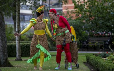 Two people in colourful costumes and hats, smiling and interacting in a garden park. One wears yellow and green, the other red, both with floral patterns, and one has a small trolley decorated with flowers.