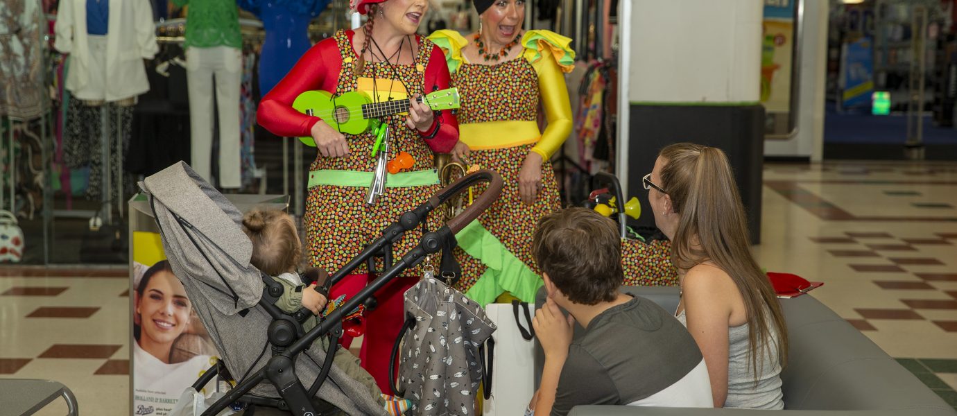 Two women in colourful costumes and flower hats sing and play a green ukulele for a seated family with three children, including one in a pushchair, inside a shopping centre. Clothing shops are visible in the background.