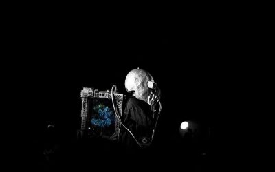 A person in a large helmet and dark clothing carries a box-like device with wires and a screen, standing in darkness with only a faint light in the background.