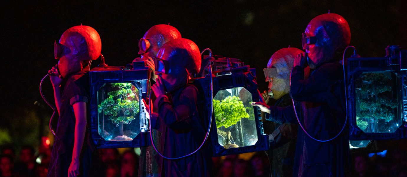 Performers in futuristic costumes with large helmets and rucksacks containing illuminated miniature trees stand in a row under dramatic red and blue lighting, with an audience watching in the background.