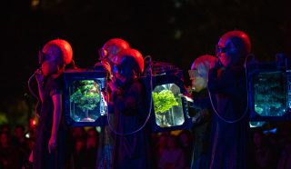 Performers in futuristic costumes with large helmets and rucksacks containing illuminated miniature trees stand in a row under dramatic red and blue lighting, with an audience watching in the background.
