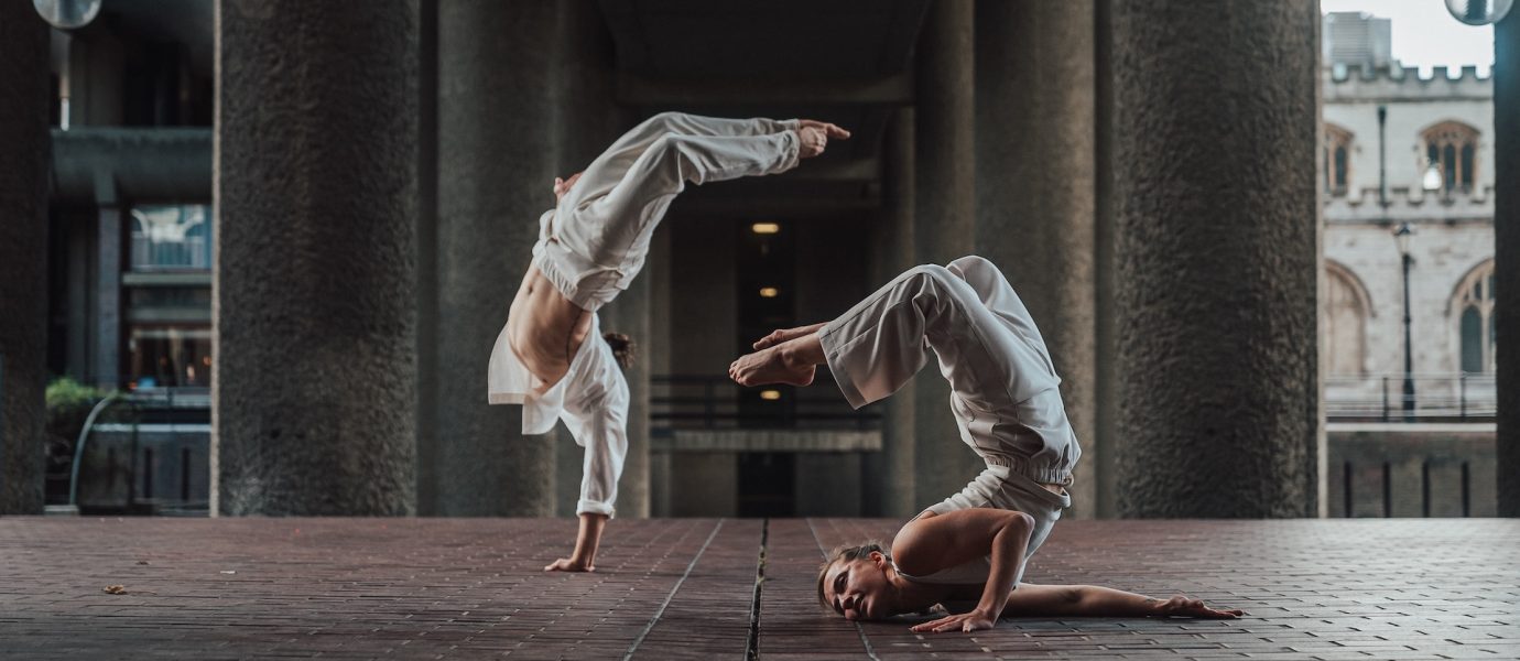 Two dancers in white outfits perform acrobatic poses on a brick floor under a concrete structure; one balances on hands, legs arched, while the other holds a headstand with legs bent, head resting on the ground.