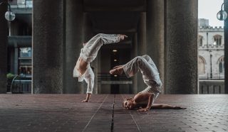 Two dancers in white outfits perform acrobatic poses on a brick floor under a concrete structure; one balances on hands, legs arched, while the other holds a headstand with legs bent, head resting on the ground.