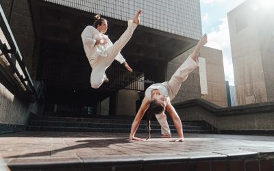 Two people in white martial arts uniforms perform dynamic capoeira moves outdoors; one jumps with a high kick whilst the other balances upside-down on their hands with a leg extended.