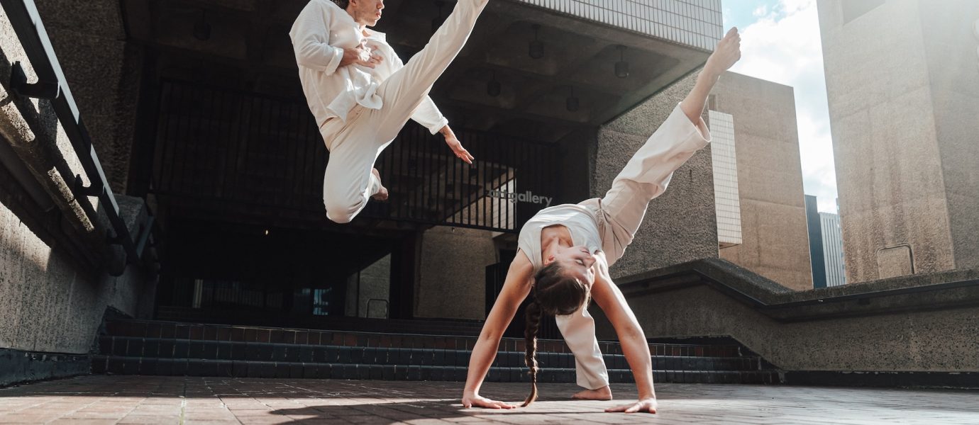 Two people in white martial arts uniforms perform dynamic moves outdoors; one is mid-air with a high kick, while the other does a one-handed handstand, both on a brick surface against modern concrete architecture.