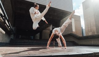 Two people in white martial arts uniforms perform dynamic moves outdoors; one jumps high with a raised leg whilst the other balances upside down on their hands, legs extended, against an urban architectural backdrop.