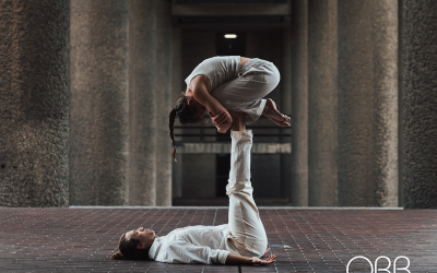 Two people practise acro yoga; one lies on their back with legs raised, supporting the other, who is balanced in a crouched position on their feet. They’re outdoors on a tiled surface, with “ORB” text in the corner.