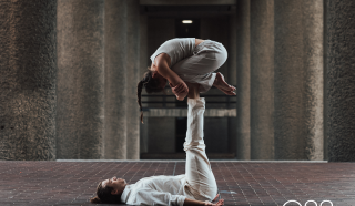 Two people practise acro yoga; one lies on their back with legs raised, supporting the other, who balances in a crouched position on their feet. Both wear white and are in an urban setting. The logo ORB appears in the corner.