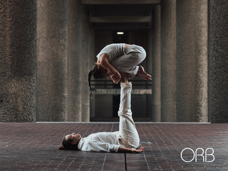 Two people practise acro yoga; one lies on their back with legs raised, supporting the other, who balances in a crouched position on their feet. Both wear white and are in an urban setting. The logo ORB appears in the corner.
