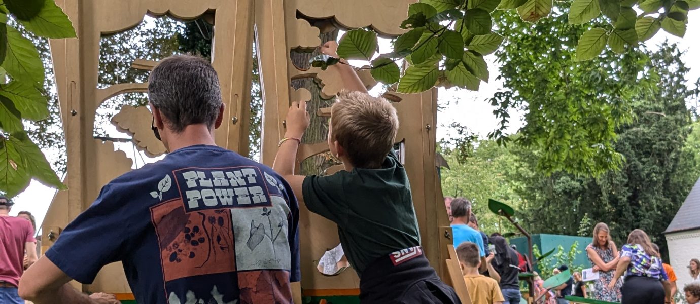 A child and an adult sit on wooden stools at outdoor game stations under trees, playing a hands-on activity at a lively park event with more people and green grass in the background.